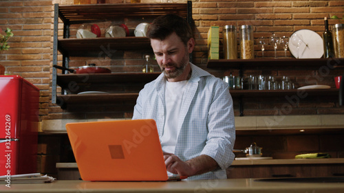 Attractive businessman working on a computer laptop in his kitchen while having breakfast. Busy man freelancing at home on a lovely day.