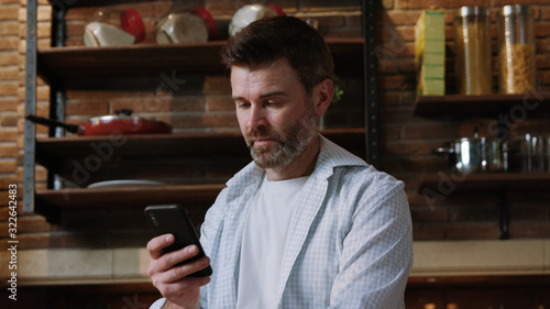 Portrait of an attractive mature man texting message on a modern smartphone standing in the middle of his kitchen in a luxury home apartment.