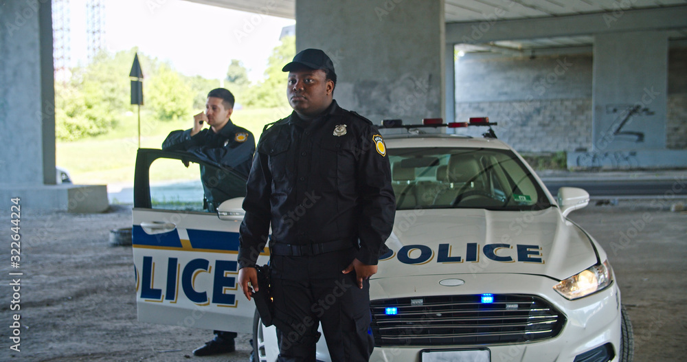 Serious handsome african american police officer looking around standing by patrol car. Two young policemen on duty keep guard on city at daytime.