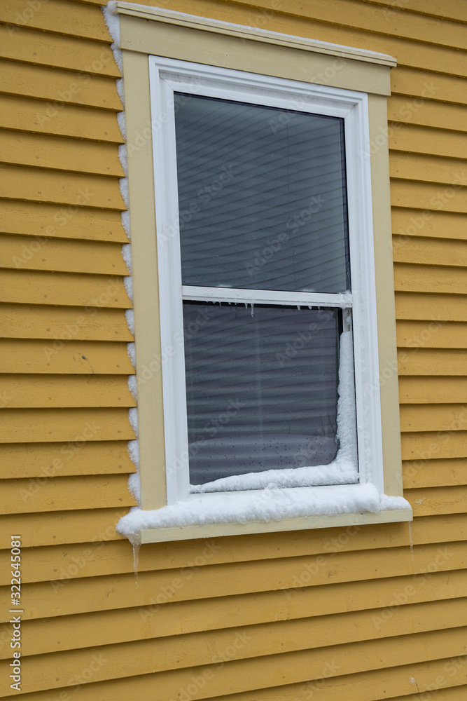 The exterior view of a double hung two pane glass window with a white mini blind. The window has beige trim around the white vinyl. The exterior wood wall of the building is a yellow in color.