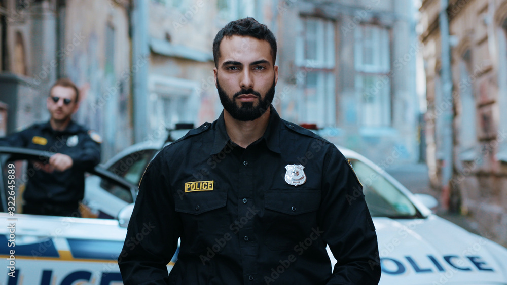 Portrait serious young man cops hold pistol stand near patrol car look at camera enforcement officer police uniform auto safety security communication control policeman portrait close up slow motion Portrait serious young man cops hold pistol stand near patrol car look at camera enforcement officer police uniform auto safety security communication control policeman portrait close up slow motion