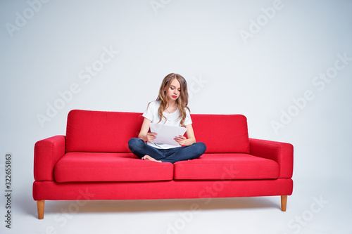 Work and relaxation. Pretty young woman reading documents while sitting on red sofa on white background. © luengo_ua