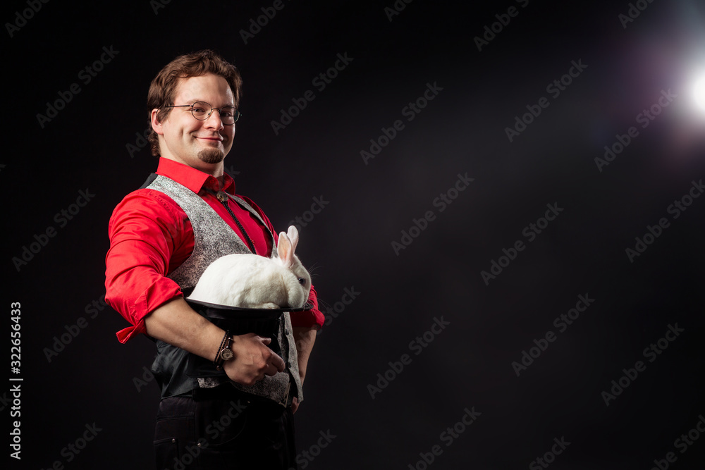 A magician is holding a cylinder hat with a white rabbit smiling. Focus miraculous appearance of extinction. Illusion wizard sleight of hand. Magic illusionist circus performance show. Funny pleased