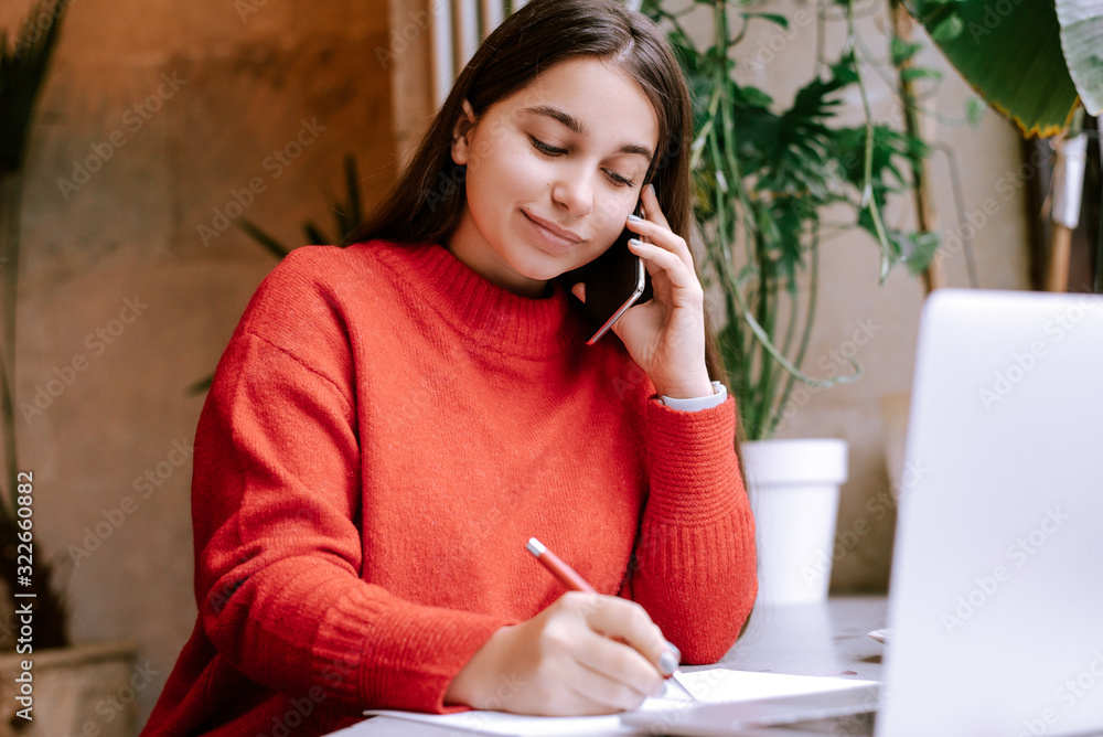 Young beautiful brunette woman in red sweater making notes in notebook and talking on phone. Female freelancer connecting to internet via computer. Blogger or journalist writing new article Young beautiful brunette woman in red sweater making notes in notebook and talking on phone. Female freelancer connecting to internet via computer. Blogger or journalist writing new article