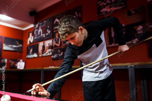 Boy plays billiard or pool in club. Young Kid learns to play snooker. Boy with billiard cue strikes the ball on table. Active Leisure, sport, hobby concept