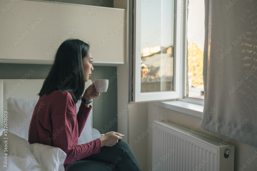 Woman is drinking hot coffee and looking out to the window in her apartment. Woman is drinking hot coffee and looking out to the window in her apartment.