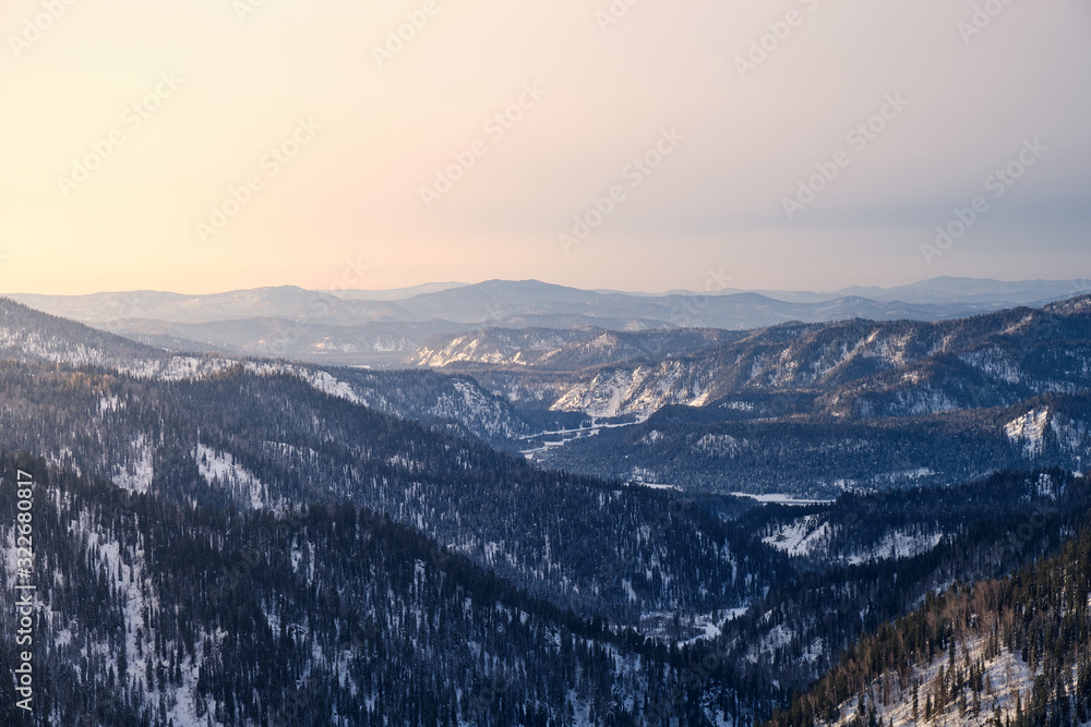 Winter mountain landscape. The view from the top. Snowy mountains at sunset, covered with coniferous forest. Russia. Altai Republic.