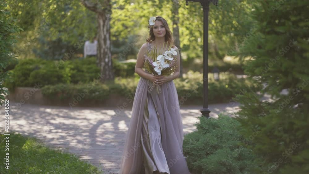 A young beautiful happy girl, a bride with a beautiful bouquet in her hands walks through the park, dressed in a colorful wedding dress, looks at the sunlight and the camera lens A young beautiful happy girl, a bride with a beautiful bouquet in her hands walks through the park, dressed in a colorful wedding dress, looks at the sunlight and the camera lens