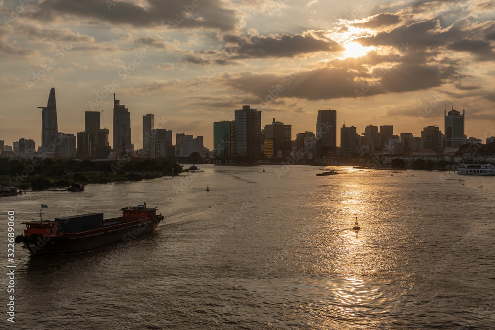 Fototapeta premium Zachód słońca nad rzeką Sajgon z panoramą miasta Ho Chi Minh o złotej godzinie. Prezentowany jest frachtowiec rzeczny, dramatyczne niebo wietnamskiej stolicy finansowej