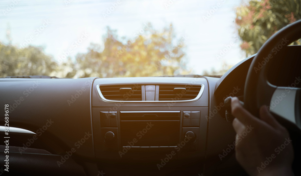Driver's hand on the black steering wheel with dashboard inside of a car and green trees against blue sky with clouds. Vintage tone.