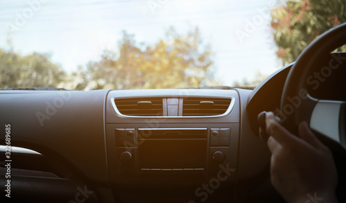 Driver's hand on the black steering wheel with dashboard inside of a car and green trees against blue sky with clouds. Vintage tone.