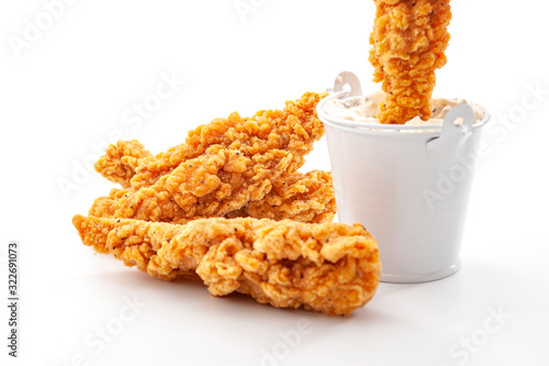 Eating fast food and protein high diet concept with golden breaded chicken strips and one crispy strip being dipped in a metal bucket of ranch dressing sauce isolated on white background photo