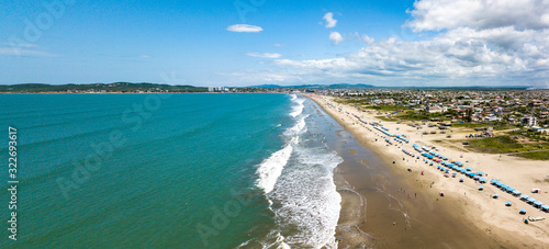 Aerial drone view of the beach, tourists, sea and waves splashing in the coast of the town Playas General Villamil, Ecuador. Sunny day. Beach houses and the horizon with some clouds in background.