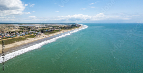Aerial drone view of the beach, tourists, sea and waves splashing in the coast of the town Playas General Villamil, Ecuador. Sunny day. Beach houses and the horizon with some clouds in background.