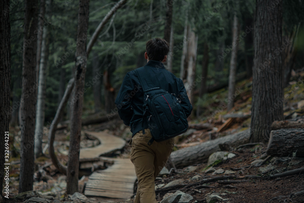 Teenager walking in the forest on wooden path with bag looking around. Adventuring wearing black jacket and beige pants.