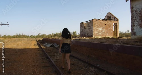 Slow motion of a young female with dark brown hair walking down the railway line of an old train station in a ghost town photo