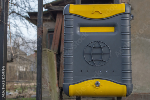 Locked plastic dark gray and yellow mailbox with globe symbol on it, with white envelope inside. Box is attached to black metal fence made of vertical rods, behind which can see dilapidated housing