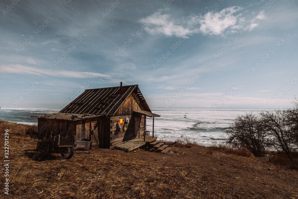 Russia. Baikal. Olkhon. An old wooden hut, the house stands on the shore. A yellow light shines through the windows. Against the background of a lake covered with ice. March, night