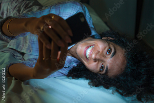 young happy and attractive hipster woman cozy on bed at night enjoying internet social media app on mobile phone online dating and smiling cheerful in cell addiction concept