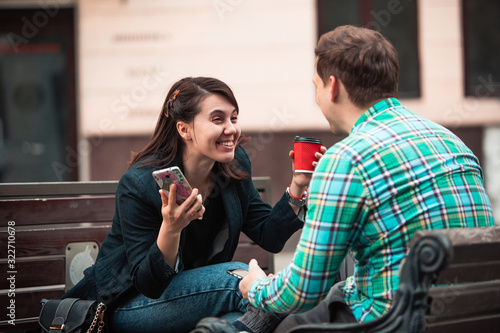 laughing talking man with woman sitting on bench drinking coffee surfing in internet. urban lifestyle