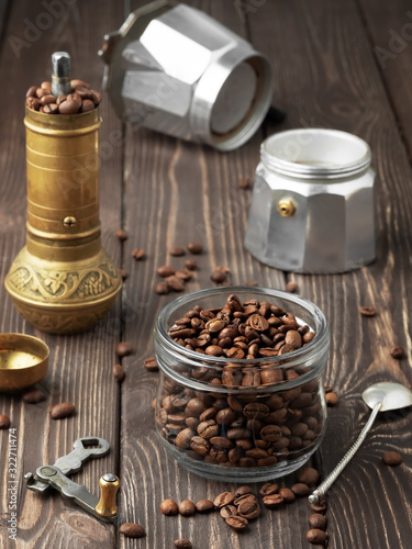Roasted coffee grains in a glass jar and scattered coffee grains on a wooden brown table. A hand mill and a coffee pot (la moka) in the background. Close Up, concept of traditional coffee preparation.