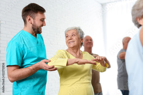Care worker helping elderly woman to do sports exercise in hospital gym.