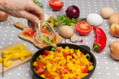 Hand pours oil. Sliced tomatoes and greens in pan. Eggs, olives, garlic, yellow pepper on table. Gray Polka Dot Background. photo