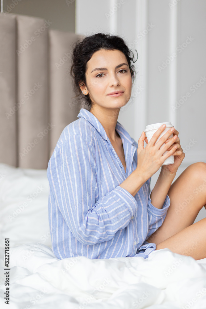 Young pretty brunette woman in pajamas shirt drinking coffee enjoy morning new day start in bed. Beautiful millennial lady looking at camera. Shot from side. Blurred bedroom interior on backdrop Young pretty brunette woman in pajamas shirt drinking coffee enjoy morning new day start in bed. Beautiful millennial lady looking at camera. Shot from side. Blurred bedroom interior on backdrop