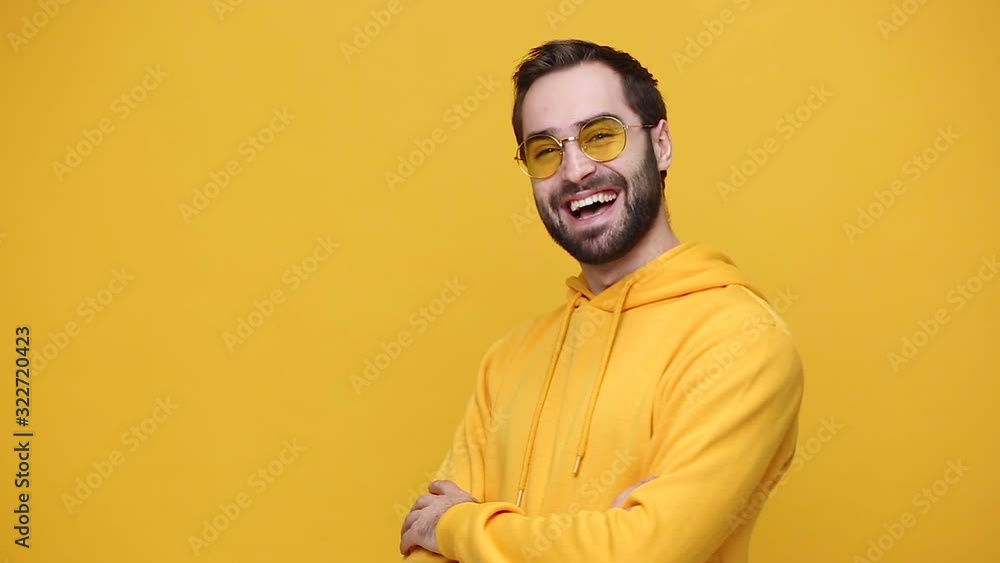 Handsome bearded young guy 20s in yellow streetwear hoodie glasses isolated on yellow background studio. People emotions, lifestyle concept. Looking aside and at camera with charming smile laughing Handsome bearded young guy 20s in yellow streetwear hoodie glasses isolated on yellow background studio. People emotions, lifestyle concept. Looking aside and at camera with charming smile laughing