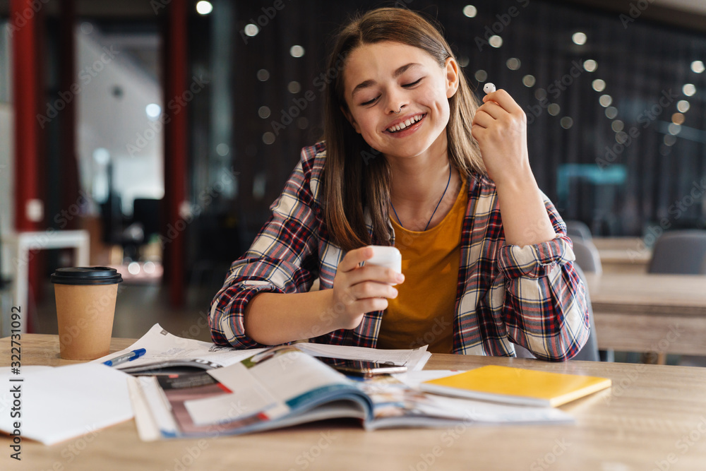 Image of happy nice girl using wireless earphone while doing homework Image of happy nice girl using wireless earphone while doing homework