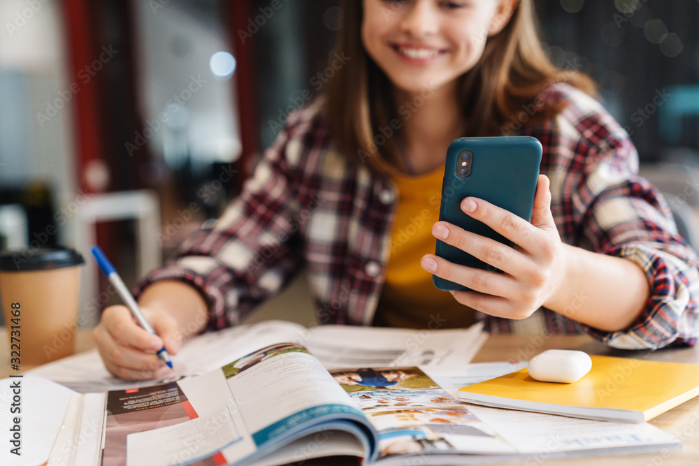 Image of joyful beautiful girl using cellphone while doing homework Image of joyful beautiful girl using cellphone while doing homework