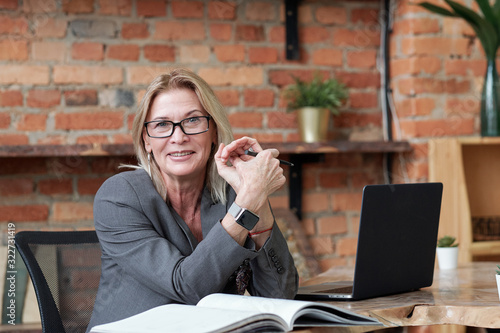 Portrait of smiling mature woman in glasses sitting at desk with laptop in loft office and examining notes in accounting book photo