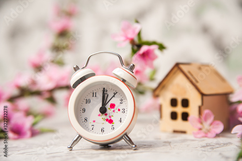Miniature clock and small wooden house close-up and copy space. Alarm clock, house, flowering branch in the spring. The concept of rush, purchase, exchange, home return and time.