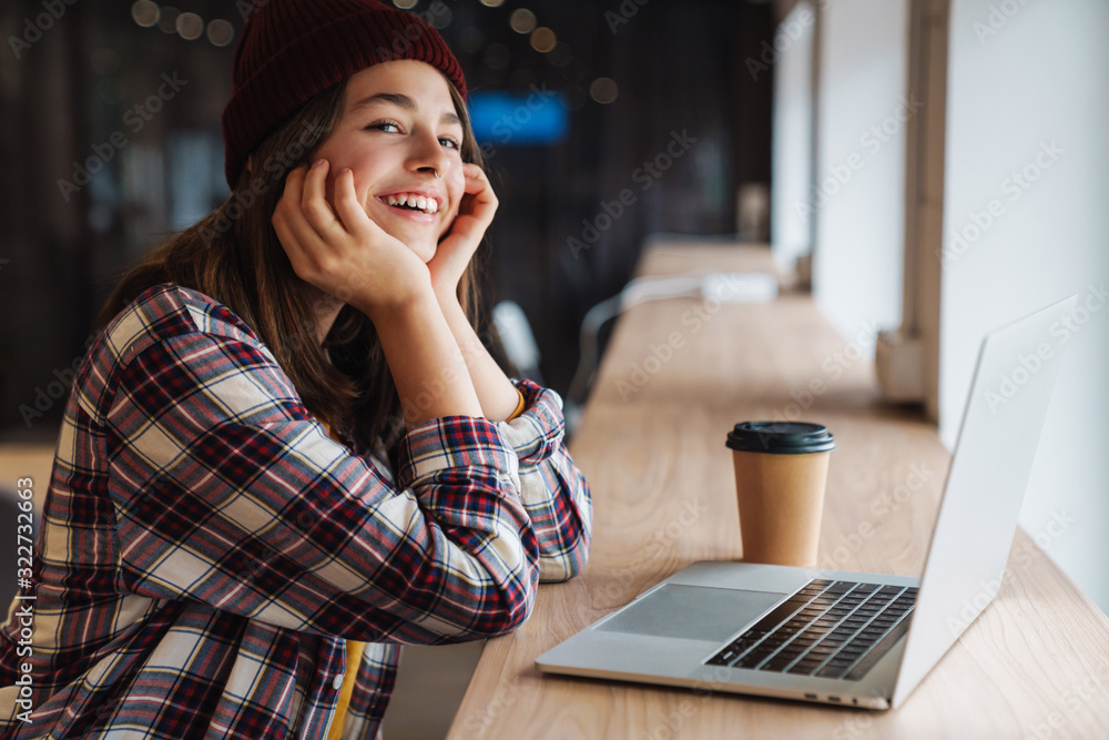 Image of joyful caucasian girl in hat drinking coffee and using laptop Image of joyful caucasian girl in hat drinking coffee and using laptop