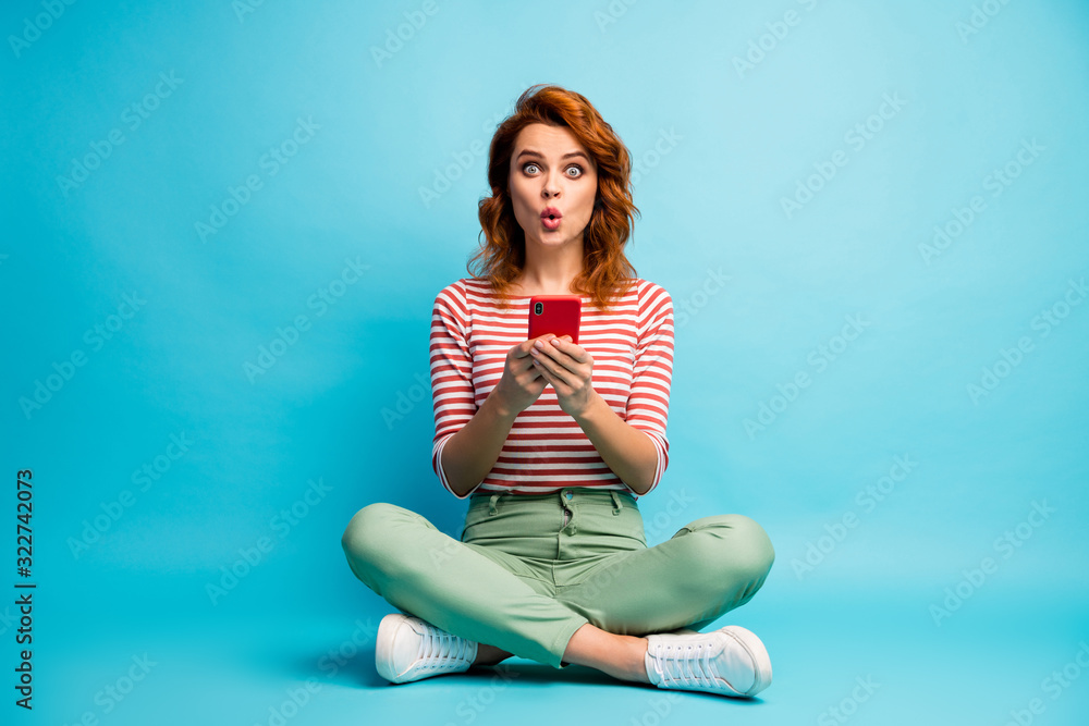 Full body photo of astonished girl sit legs crossed use cell phone read social media information impressed scream wow omg wear stylish clothing isolated over blue color background Full body photo of astonished girl sit legs crossed use cell phone read social media information impressed scream wow omg wear stylish clothing isolated over blue color background