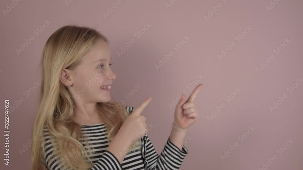 pretty smiling blonde little girl points to something posing on light pink background in light studio closeup pretty smiling blonde little girl points to something posing on light pink background in light studio closeup