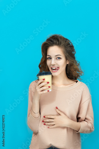 Portrait of a young beautiful woman wearing sweatshirt holding a cup with hot drink in hand and stroking belly looking at you over blue background