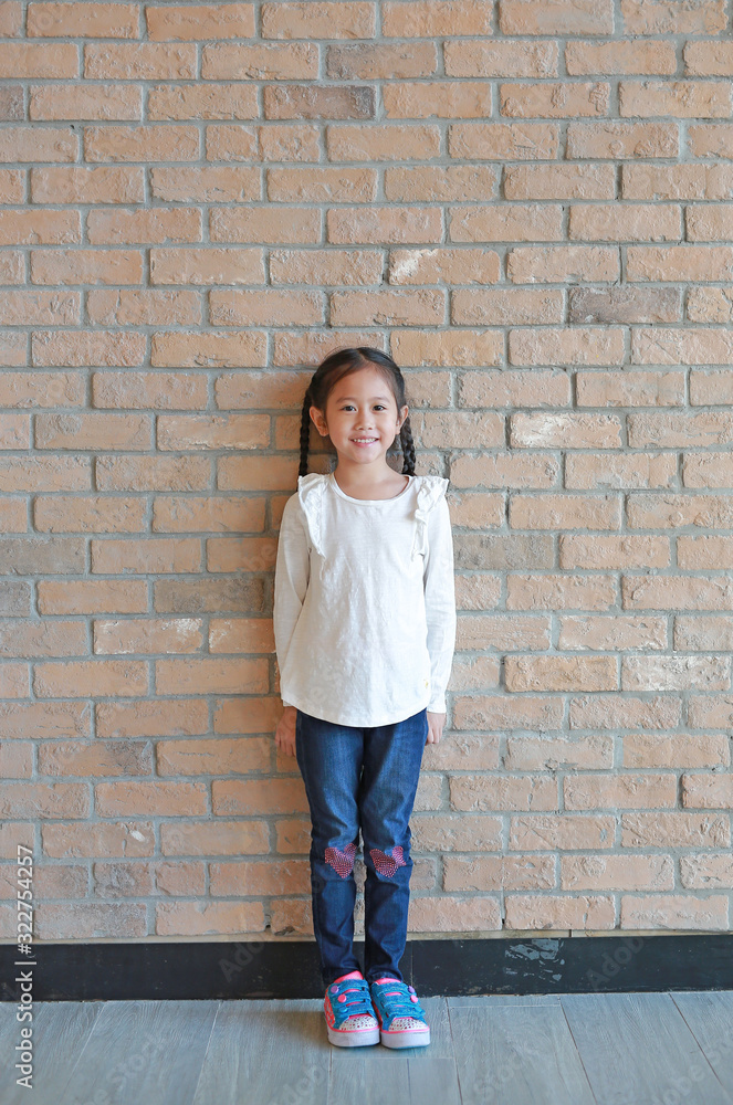 Portrait of happy asian little girl with pigtail hair in jeans and a white shirt standing at brick wall background Portrait of happy asian little girl with pigtail hair in jeans and a white shirt standing at brick wall background