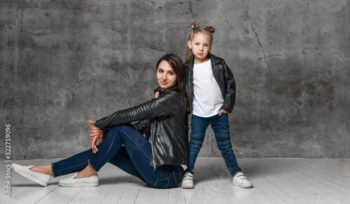 Positive young woman and little girl in similar clothing jeans and black leather jackets posing over grey concrete background