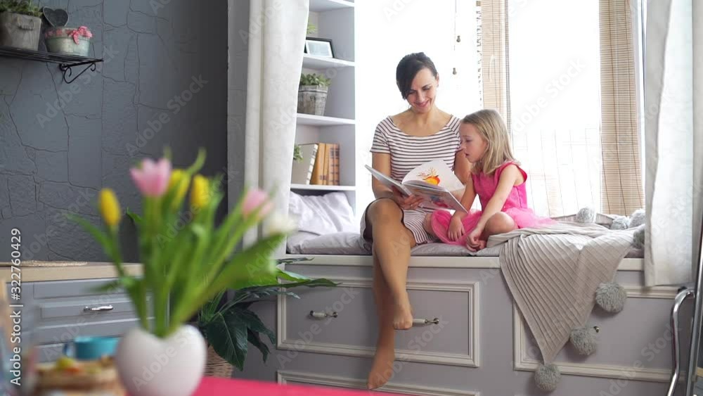Young Woman Reading Book with Little Daughter at Home. They Sitting on the Big Cozy Windowsill. Slow Motion. People, Lifestyle and Happy Family Concept Young Woman Reading Book with Little Daughter at Home. They Sitting on the Big Cozy Windowsill. Slow Motion. People, Lifestyle and Happy Family Concept