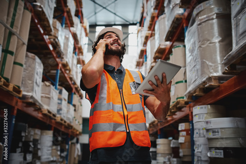 Low angle view of warehouse manager talking over phone while smiling and holding digital tablet standing in aisle with goods