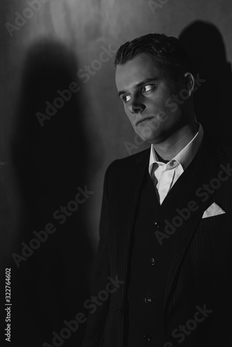 Retro fashion man in dark suit with angry look stands against wall.