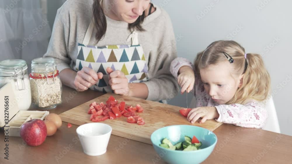 Mom teaches to cook a little daughter. Girl tries to cut tomato for salad Mom teaches to cook a little daughter. Girl tries to cut tomato for salad