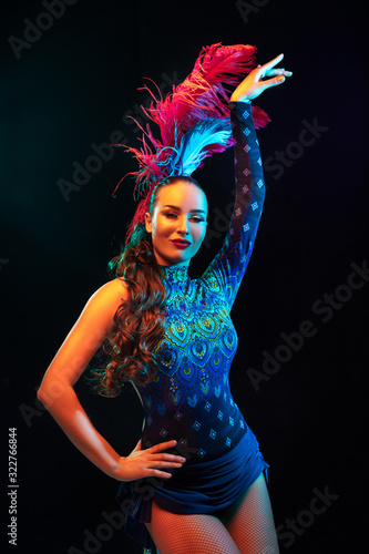 Mystically. Beautiful young woman in carnival, stylish masquerade costume with feathers on black background in neon light. Copyspace for ad. Holidays celebration, dancing, fashion. Festive time, party