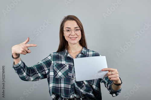 Young brunette girl blue green in checked shirt on grey background a happy woman with glasses holds an empty white sheet of paper in hand, points at it with index finger