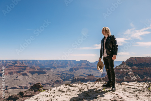 A curly haired blonde man, wearing surprisingly a black leather jacket ,black ripped jeans, black shoes, and a beige linen shirt, is tipping his cowboy hat to the majestic beauty of the Grand Canyon