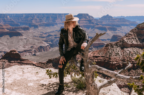 A Curly haired blonde man, wearing a black leather jacket ,black jeans, black shoes, beige linen shirt and matching cowboy hat, perched on an old tree stump with background vistas of the Grand Canyon