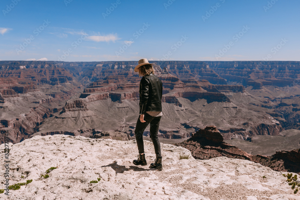 A Curly haired blonde man, wearing a black leather jacket ,black jeans, black shoes, beige linen shirt and matching cowboy hat, background vistas of the Grand Canyon