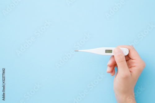 Young woman hands holding white digital thermometer on pastel blue background. Fever and healthcare concept. Closeup. Point of view shot. Empty place for text. Top down view. Copy space.