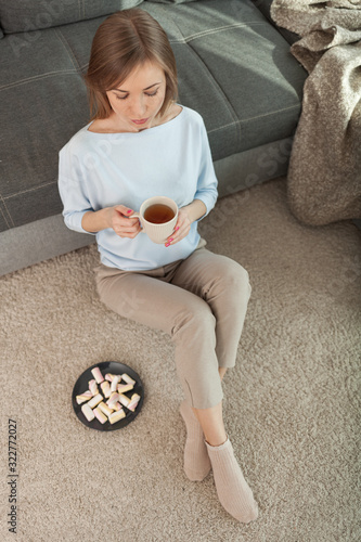 Young woman holds cup of tea, sitting on the carpet, relaxing at the day in apartments. Top view